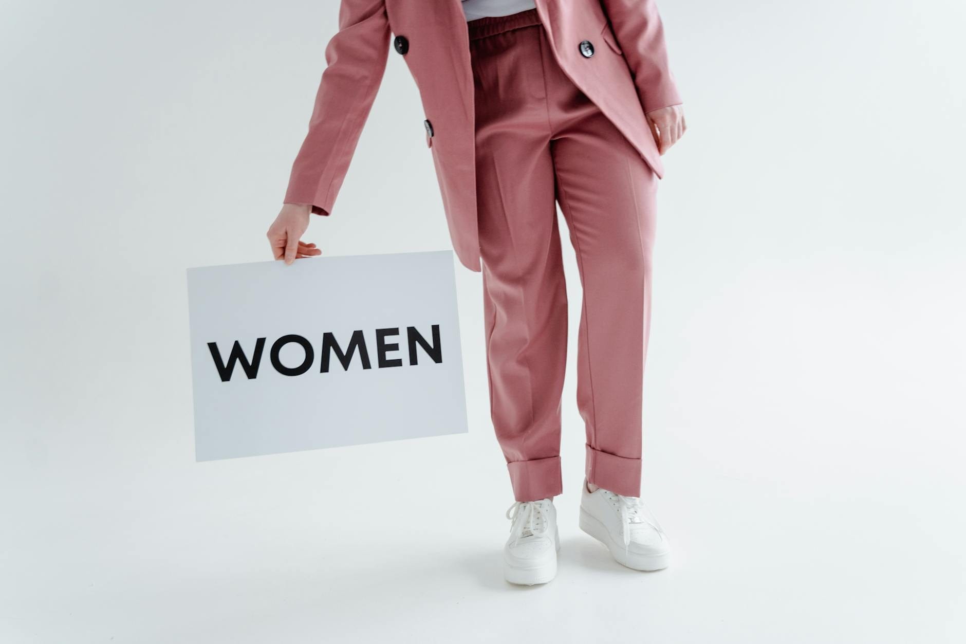 Kobieta w różowym garniturze trzyma kartkę z napisem WOMEN